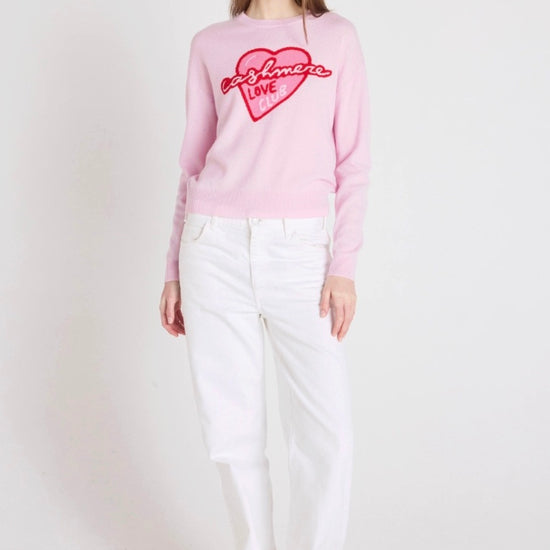 Woman wearing a pink sweater with a heart design and white pants on a white background