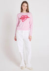 Woman wearing a pink sweater with a heart design and white pants on a white background
