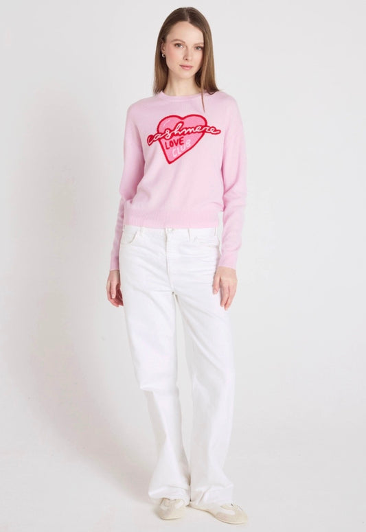 Woman wearing a pink sweater with a heart design and white pants on a white background
