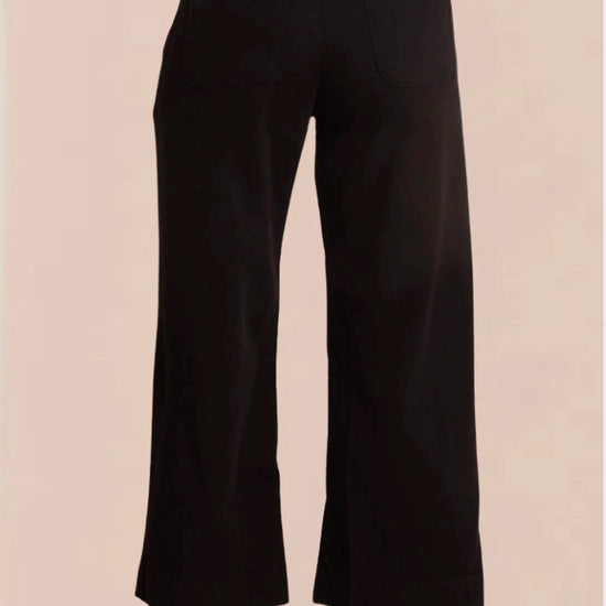 A female model wearing black wide trousers with a fixed waistband and a white, short-sleeved, button-up top. Accessory details include black heels.