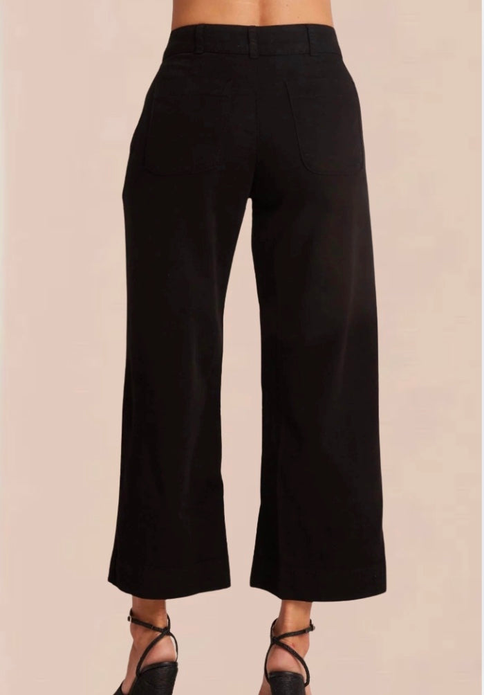 A female model wearing black wide trousers with a fixed waistband and a white, short-sleeved, button-up top. Accessory details include black heels.