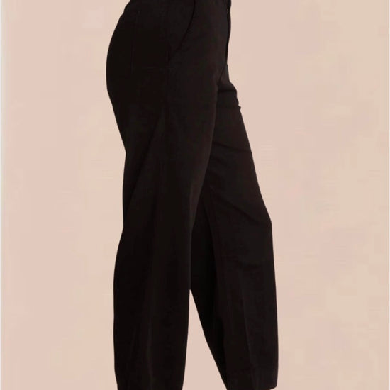 A female model wearing black wide trousers with a fixed waistband and a white, short-sleeved, button-up top. Accessory details include black heels.