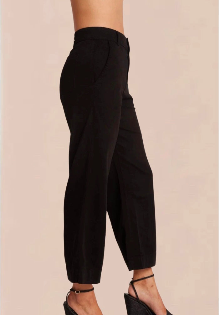 A female model wearing black wide trousers with a fixed waistband and a white, short-sleeved, button-up top. Accessory details include black heels.