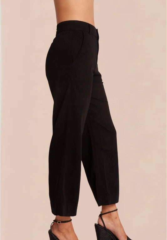 A female model wearing black wide trousers with a fixed waistband and a white, short-sleeved, button-up top. Accessory details include black heels.