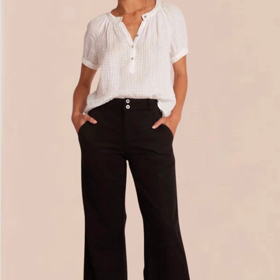 A female model wearing black wide trousers with a fixed waistband and a white, short-sleeved, button-up top. Accessory details include black heels.
