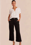 A female model wearing black wide trousers with a fixed waistband and a white, short-sleeved, button-up top. Accessory details include black heels.