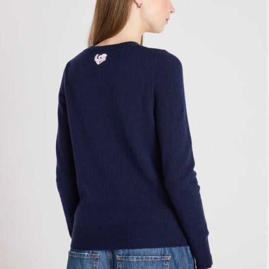 Woman wearing a navy sweater with a heart design on the back, standing against a light gray background.