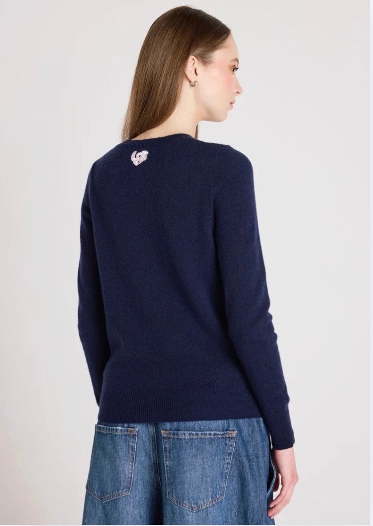 Woman wearing a navy sweater with a heart design on the back, standing against a light gray background.