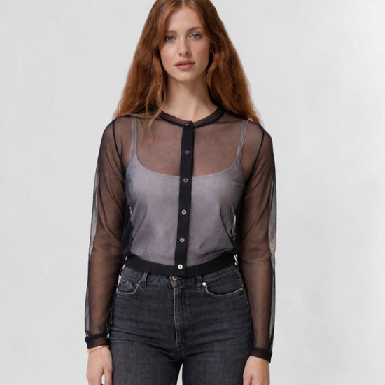 Woman wearing a sheer black blouse over a gray top with dark jeans on a white background