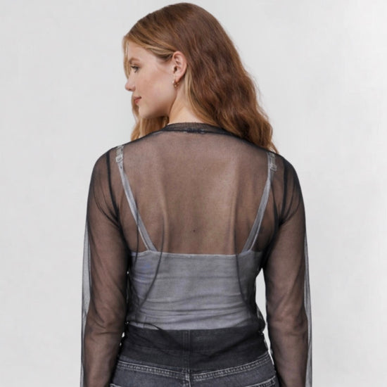 Woman wearing a sheer long-sleeve top over a gray shirt with dark jeans on a white background