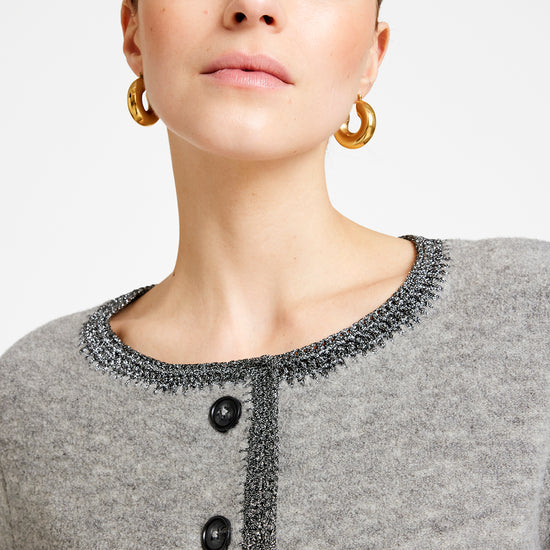 Woman wearing gold hoop earrings and a gray cardigan with black buttons on a light background