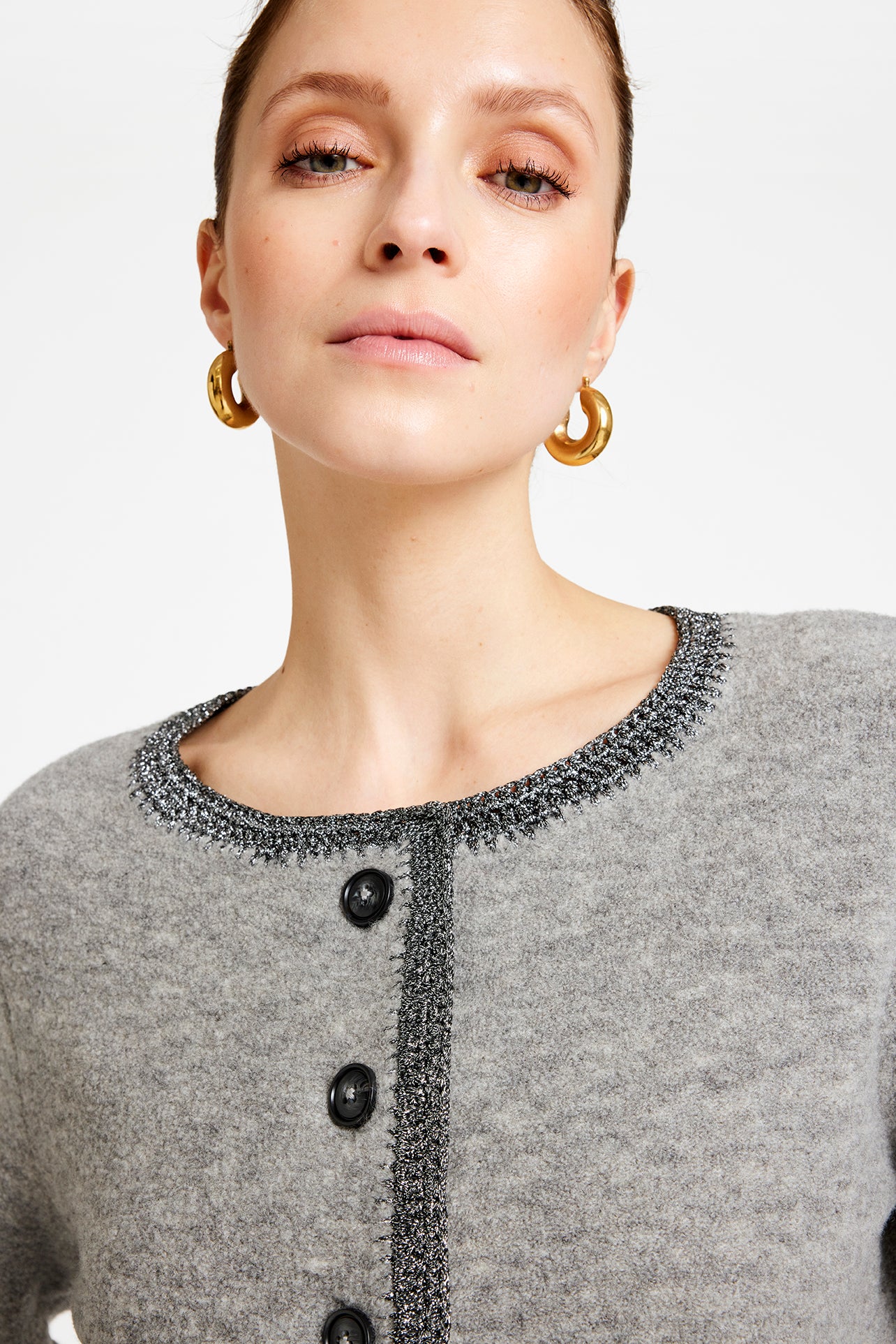 Woman wearing gold hoop earrings and a gray cardigan with black buttons on a light background