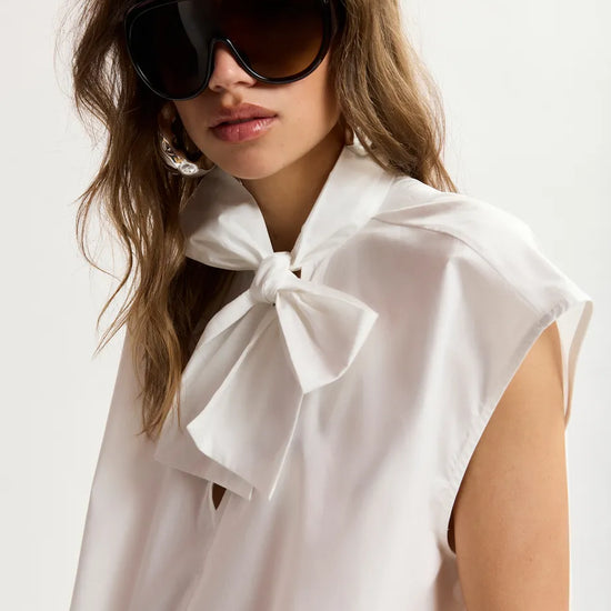 Woman wearing a white blouse with a bow and sunglasses on a plain background
