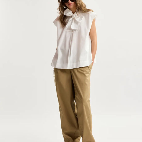Woman wearing a white sleeveless top with a bow and beige pants on a white background