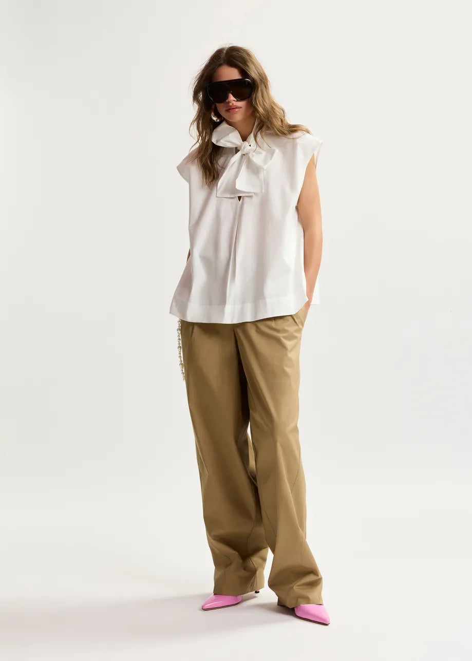 Woman wearing a white sleeveless top with a bow and beige pants on a white background