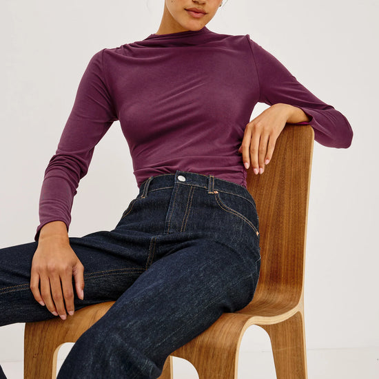 A woman wearing a fitted long-sleeve mock neck top in deep plum purple and dark blue jeans, sitting on a wooden chair against a plain light background.