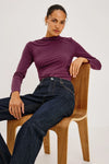 A woman wearing a fitted long-sleeve mock neck top in deep plum purple and dark blue jeans, sitting on a wooden chair against a plain light background.