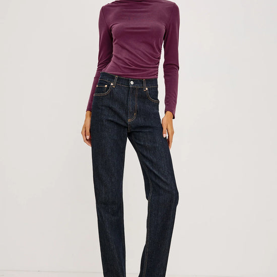 A woman wearing a fitted long-sleeve mock neck top in deep plum purple and dark blue jeans, standing full length against a light grey background