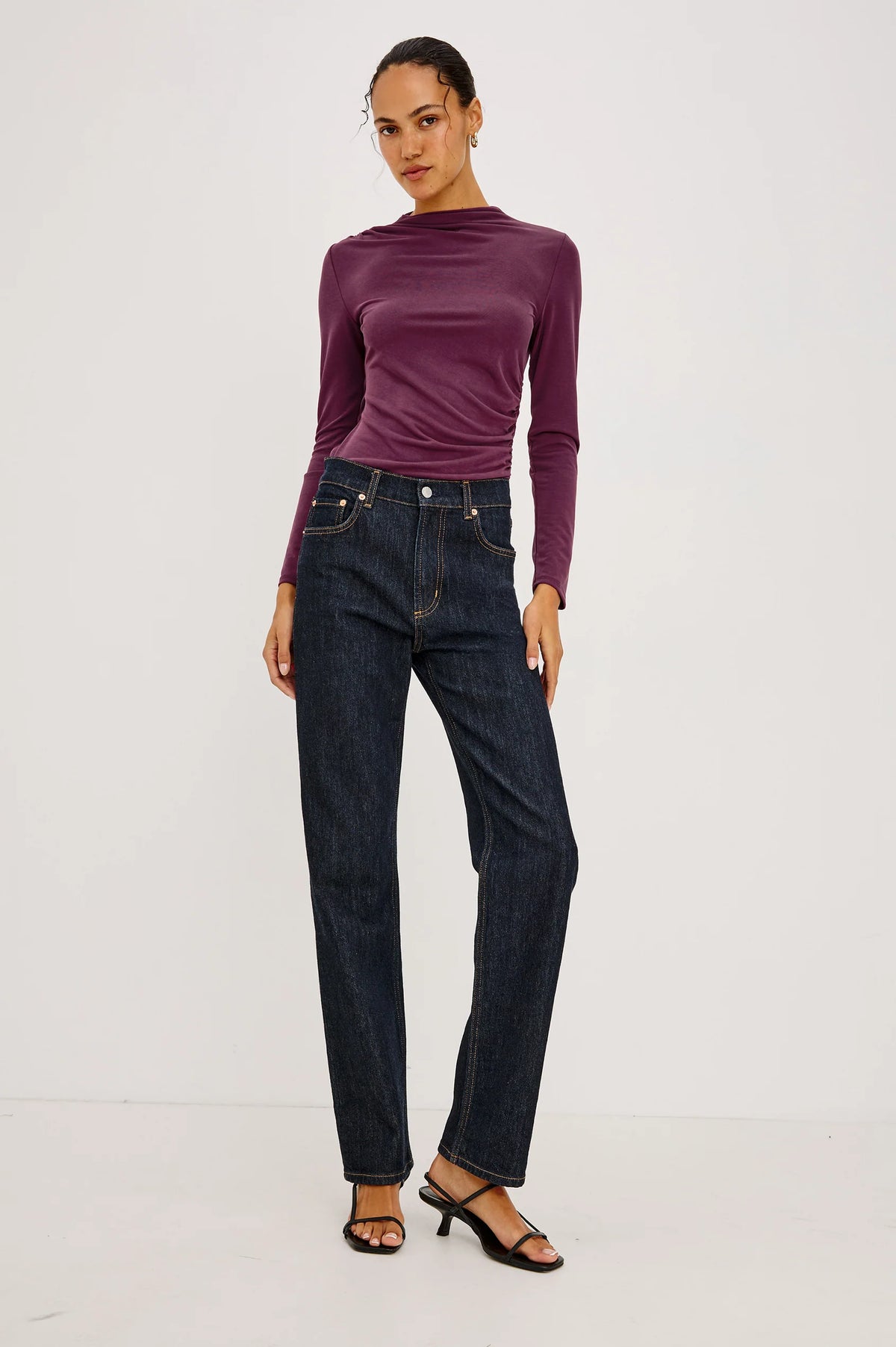 A woman wearing a fitted long-sleeve mock neck top in deep plum purple and dark blue jeans, standing full length against a light grey background