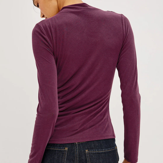 A woman shown from the back wearing a fitted long-sleeve knit top in a deep plum purple, paired with dark blue jeans, against a plain light gray studio background.