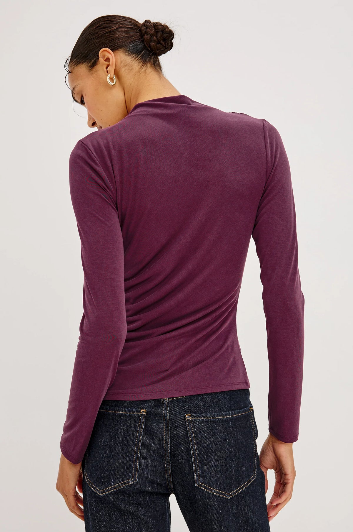 A woman shown from the back wearing a fitted long-sleeve knit top in a deep plum purple, paired with dark blue jeans, against a plain light gray studio background.