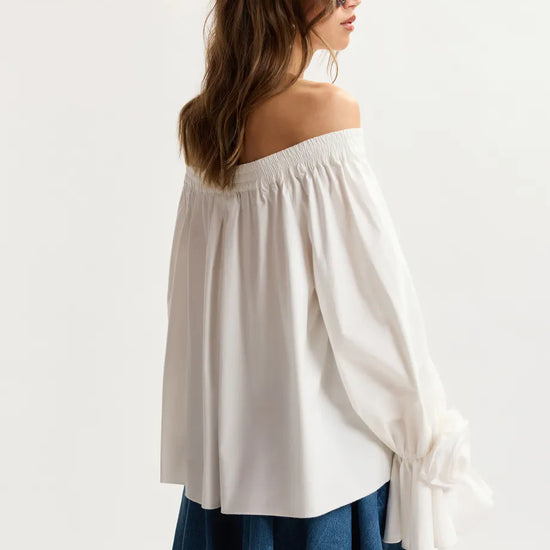 Woman wearing a white off-shoulder top and denim skirt on a plain background