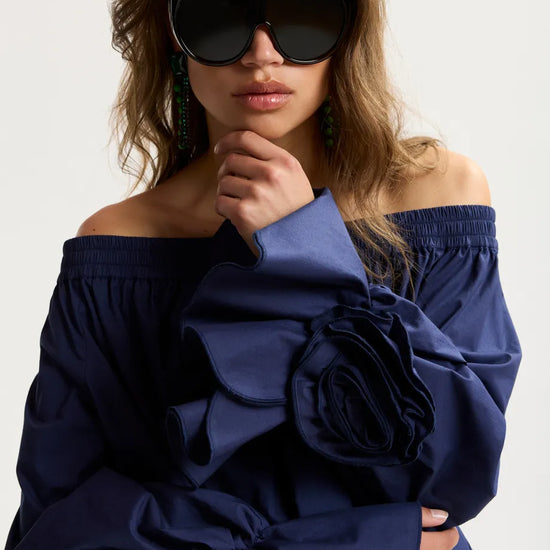 Woman wearing a blue off-shoulder dress with a large floral detail, posing against a plain background.