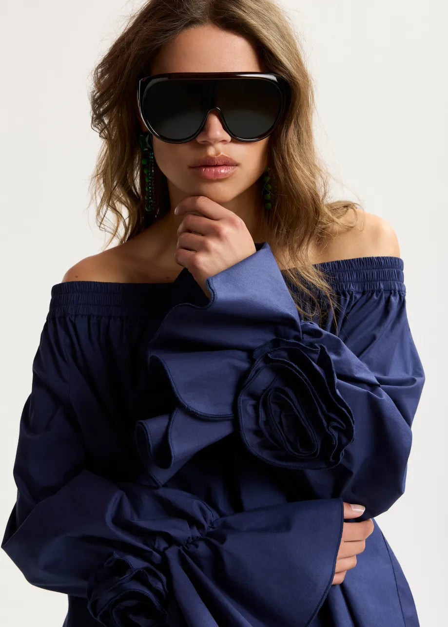 Woman wearing a blue off-shoulder dress with a large floral detail, posing against a plain background.