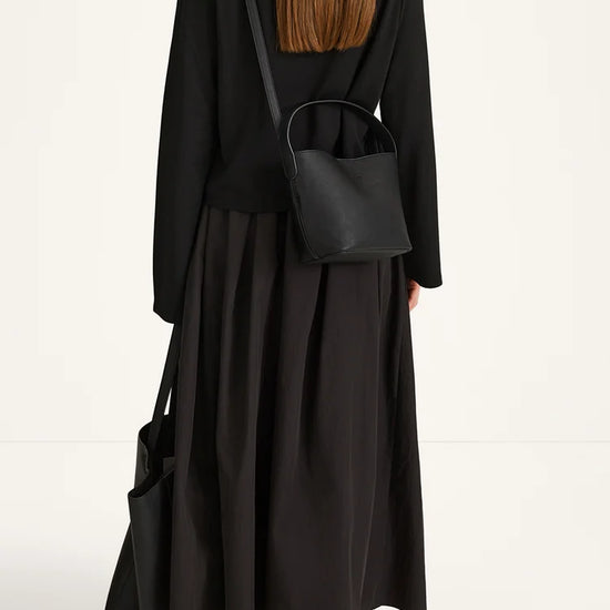 A woman shown from the back wearing a black long-sleeve top and black maxi skirt, carrying a small black leather shoulder bag and a large black tote bag, against a plain white studio background.