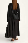 A woman shown from the back wearing a black long-sleeve top and black maxi skirt, carrying a small black leather shoulder bag and a large black tote bag, against a plain white studio background.