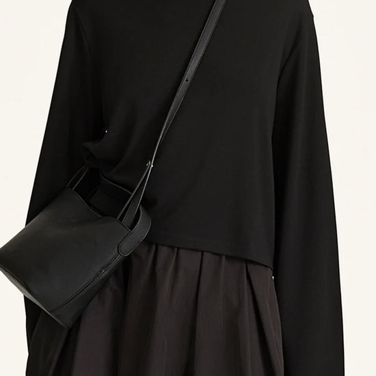 A model wearing a black long-sleeve top and black pleated skirt, carrying a black leather shoulder bag with a thin strap.