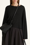 A model wearing a black long-sleeve top and black pleated skirt, carrying a black leather shoulder bag with a thin strap.