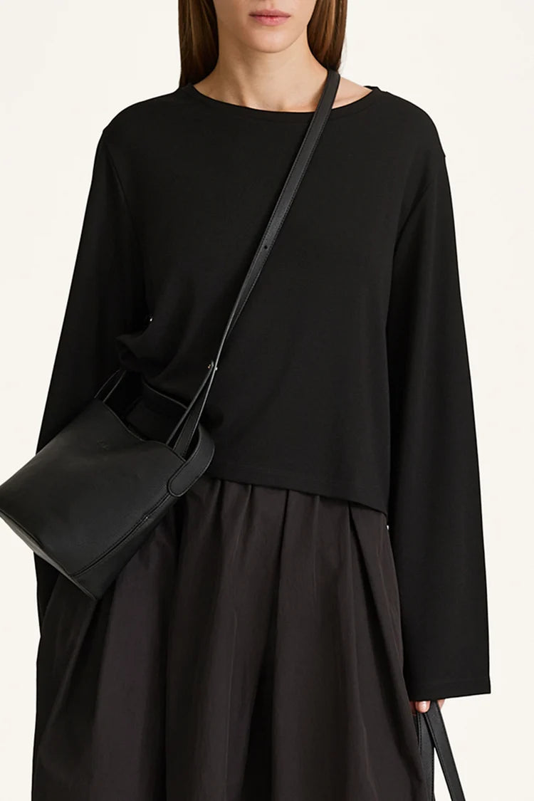 A model wearing a black long-sleeve top and black pleated skirt, carrying a black leather shoulder bag with a thin strap.