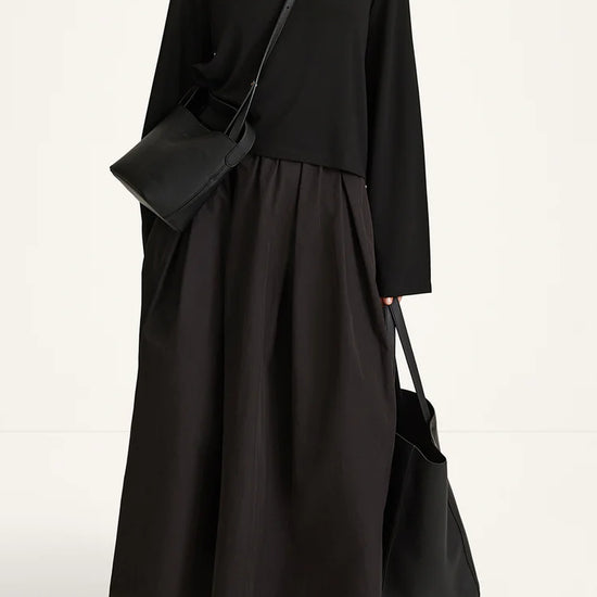 A model wearing a black long-sleeve maxi dress with a pleated skirt, styled with a black crossbody bag and a black tote bag, against a plain white studio background.