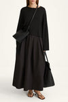 A model wearing a black long-sleeve maxi dress with a pleated skirt, styled with a black crossbody bag and a black tote bag, against a plain white studio background.