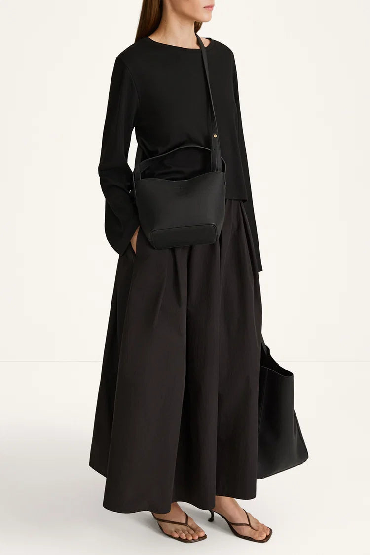A model wearing a black long-sleeve maxi dress with a matching black crossbody bag and brown strappy sandals, photographed against a clean white studio background.