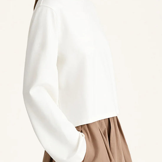 A woman wearing a white long-sleeve cropped top with a crew neck and a brown pleated skirt, shown from a side profile against a plain off-white background.