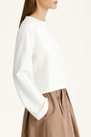 A woman wearing a white long-sleeve cropped top with a crew neck and a brown pleated skirt, shown from a side profile against a plain off-white background.
