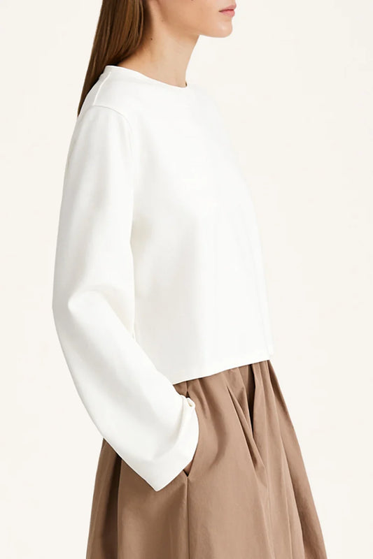 A woman wearing a white long-sleeve cropped top with a crew neck and a brown pleated skirt, shown from a side profile against a plain off-white background.
