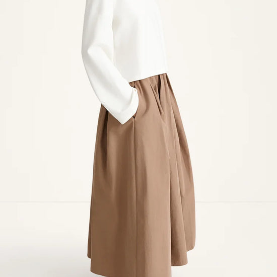 A model shown from the side wearing a white long-sleeve top and a high-waisted, pleated taupe-brown midi skirt with side pockets, paired with dark brown flat shoes, against a plain off-white studio background.