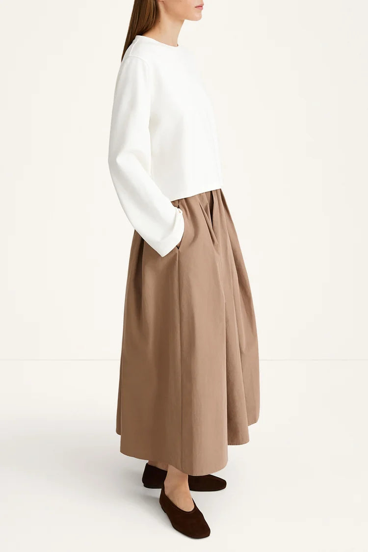 A model shown from the side wearing a white long-sleeve top and a high-waisted, pleated taupe-brown midi skirt with side pockets, paired with dark brown flat shoes, against a plain off-white studio background.