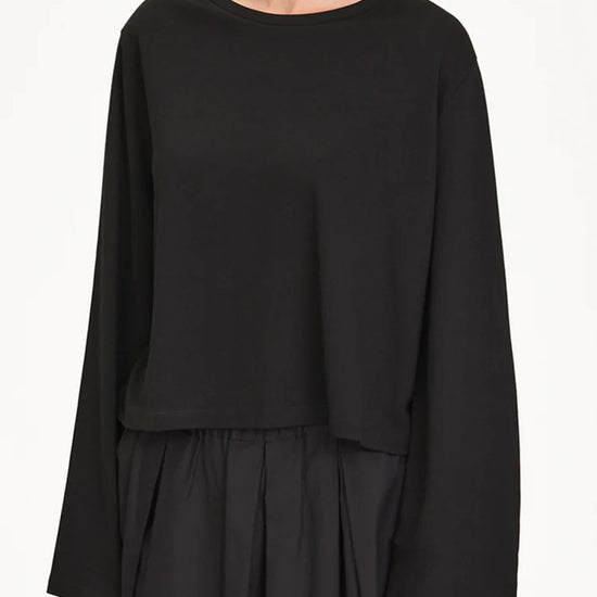 A woman wearing a black long-sleeve cropped top with a wide boat neckline and a black pleated skirt, shown against a plain white studio background.