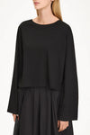 A woman wearing a black long-sleeve cropped top with a wide boat neckline and a black pleated skirt, shown against a plain white studio background.