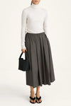 Person wearing a white turtleneck and gray skirt holding a black handbag on a white background