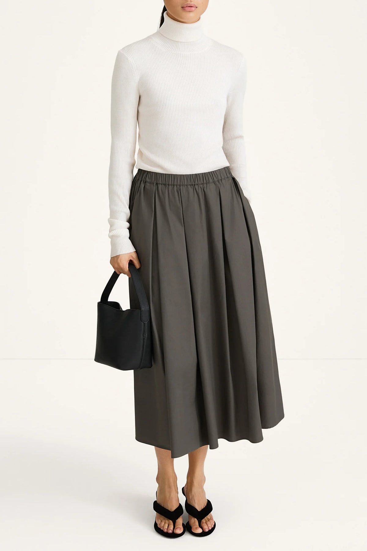 Person wearing a white turtleneck and gray skirt holding a black handbag on a white background
