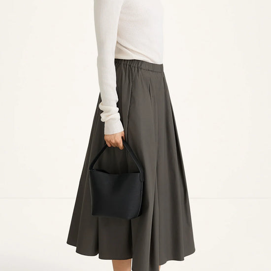 A model is shown from the side wearing a white long-sleeve turtleneck sweater, a dark gray pleated midi skirt, and black heeled sandals, while holding a black bucket bag against a plain white studio background.
