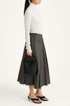 A model is shown from the side wearing a white long-sleeve turtleneck sweater, a dark gray pleated midi skirt, and black heeled sandals, while holding a black bucket bag against a plain white studio background.