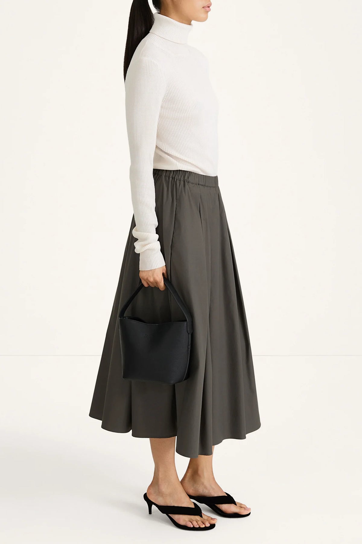 A model is shown from the side wearing a white long-sleeve turtleneck sweater, a dark gray pleated midi skirt, and black heeled sandals, while holding a black bucket bag against a plain white studio background.