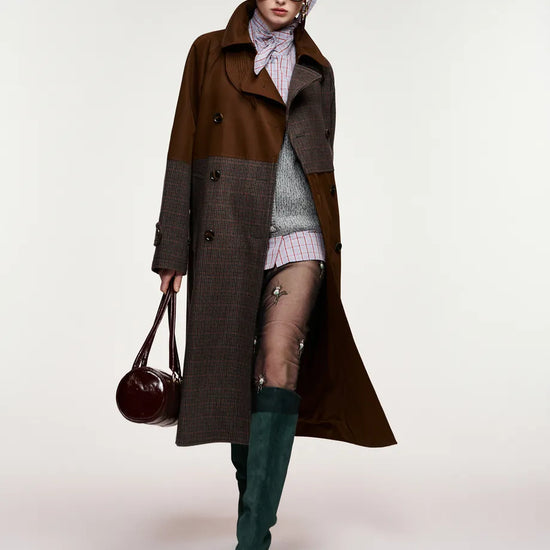 A model wearing a long brown and gray patchwork coat, a striped knit top, sheer tights, dark green knee-high boots, and carrying a dark brown leather handbag against a plain light background.