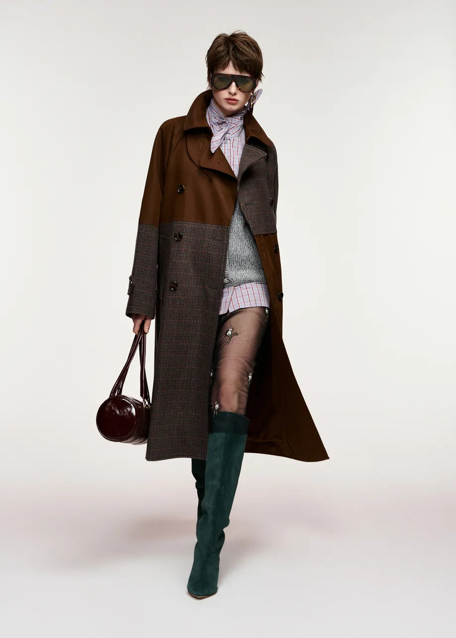 A model wearing a long brown and gray patchwork coat, a striped knit top, sheer tights, dark green knee-high boots, and carrying a dark brown leather handbag against a plain light background.